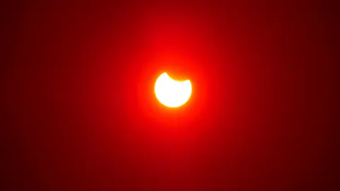 Le Soleil partiellement éclipsé : un croissant blanc mordu par la Lune, dans un ciel rouge de fin de journée.