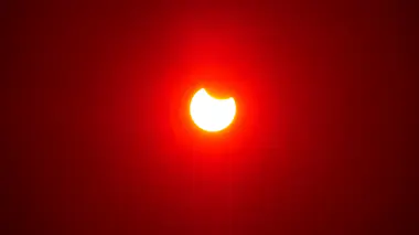 Le Soleil partiellement éclipsé : un croissant blanc mordu par la Lune, dans un ciel rouge de fin de journée.